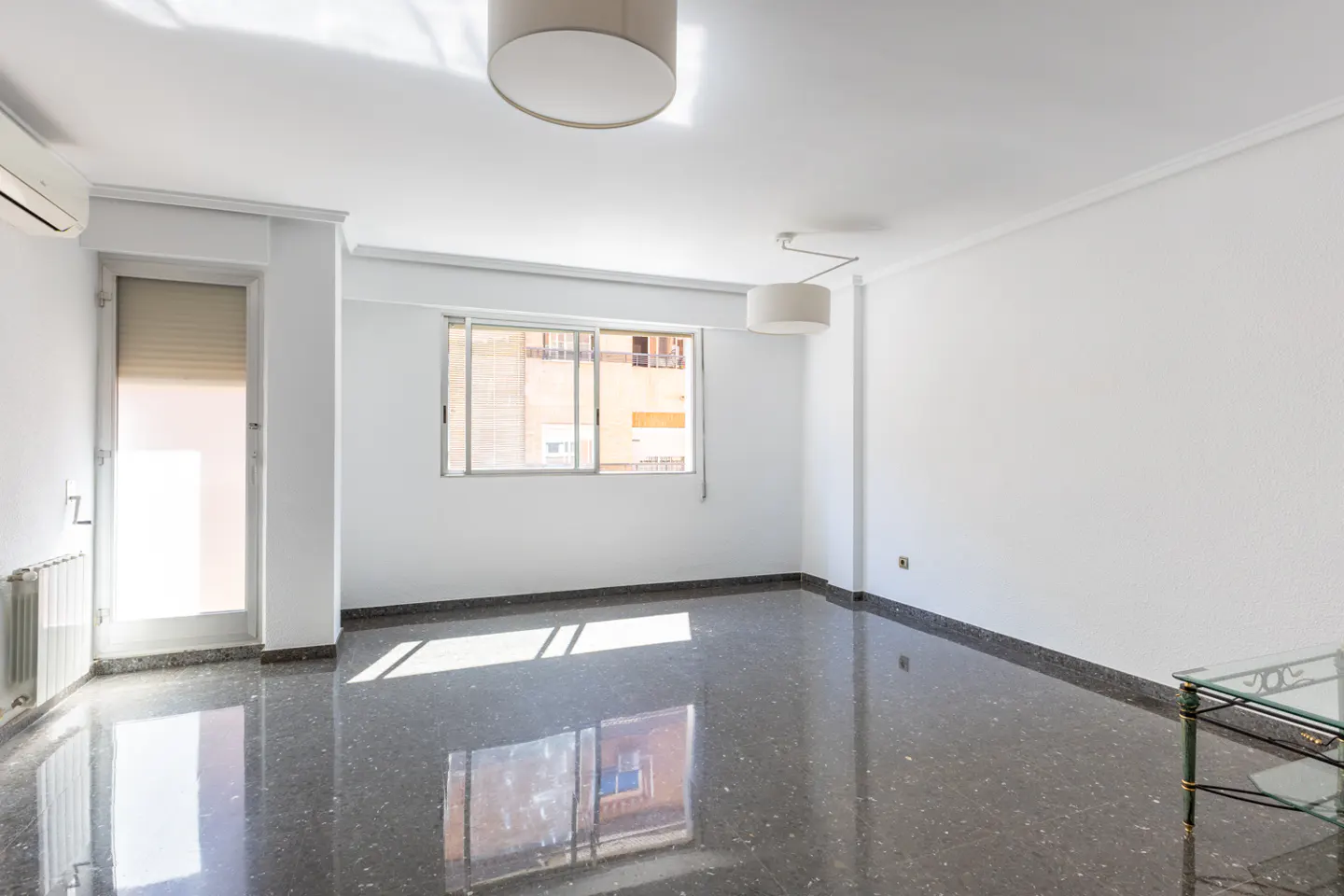 Bright, empty room with white walls, dark speckled floor, and natural light from windows. Two ceiling lights and a radiator are visible.