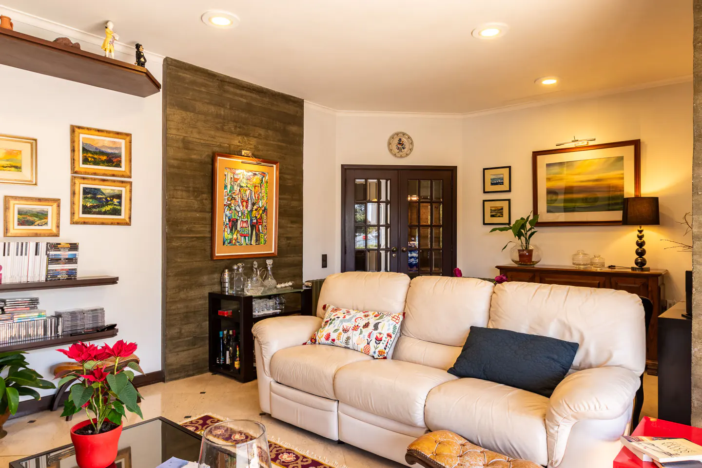 Living room with a cream sofa, dark wood accents, and artwork. Shelves display books and decor. A poinsettia adds a touch of red.