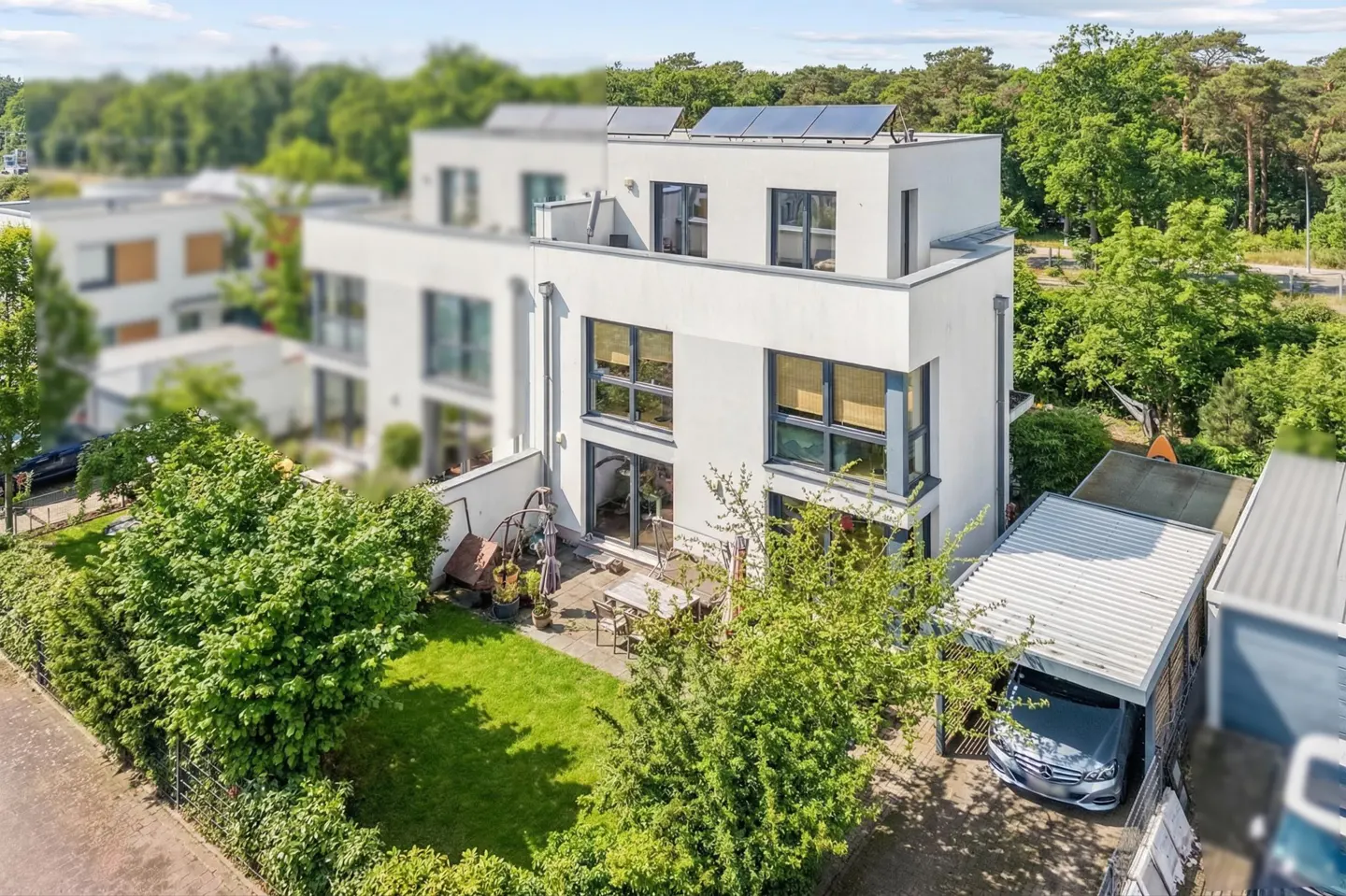 Aerial view of a modern, three-story white house with solar panels, a green lawn, patio furniture, and a carport with a car.