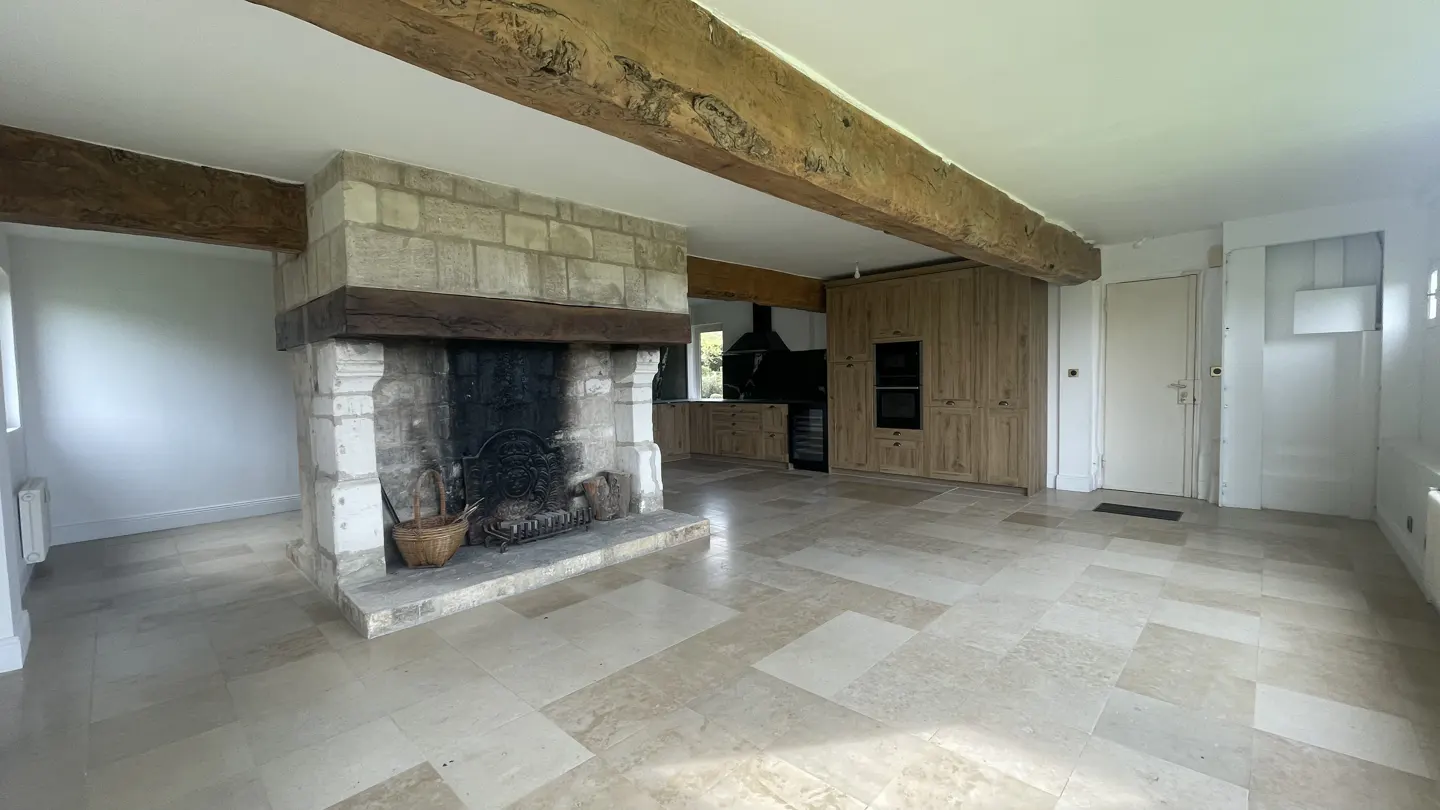 A bright, open-plan living space with a stone fireplace, wooden beams, and a view into the kitchen.
