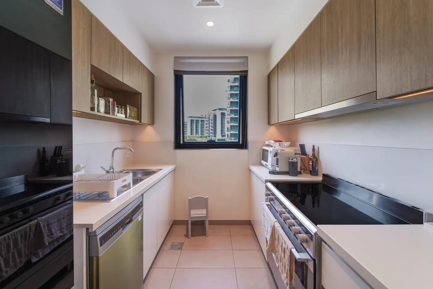 A modern kitchen with light wood cabinets, white countertops, and stainless steel appliances. A window overlooks city buildings.