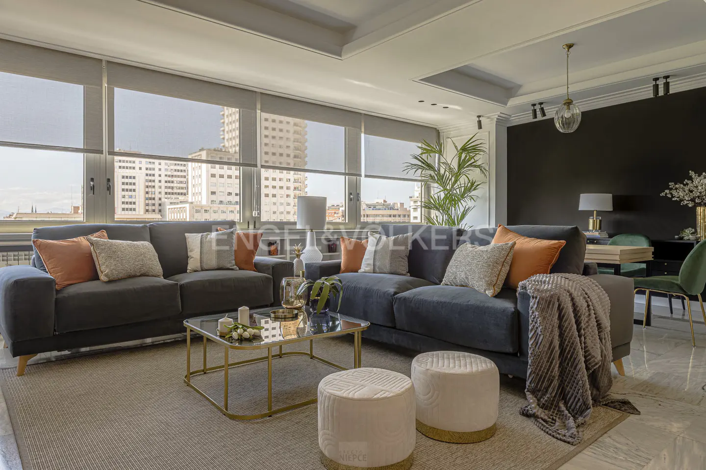 Living room with two gray sofas, pillows, coffee table, and two white ottomans. Large windows with city views.
