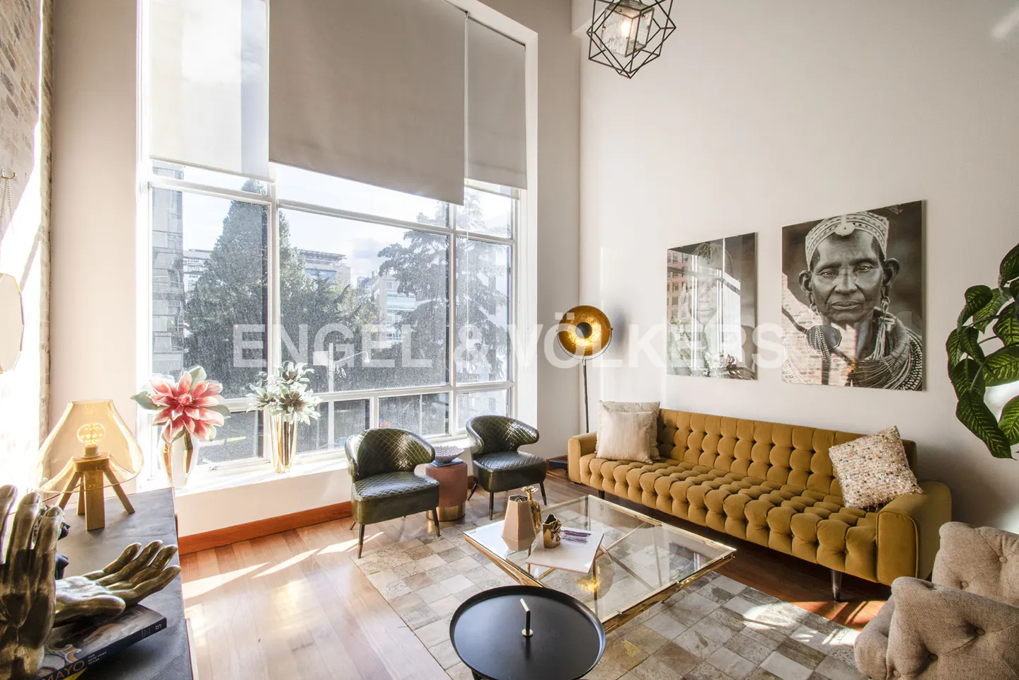 Living room with a mustard sofa, two green chairs, and large windows overlooking a cityscape. Artwork adorns the walls.