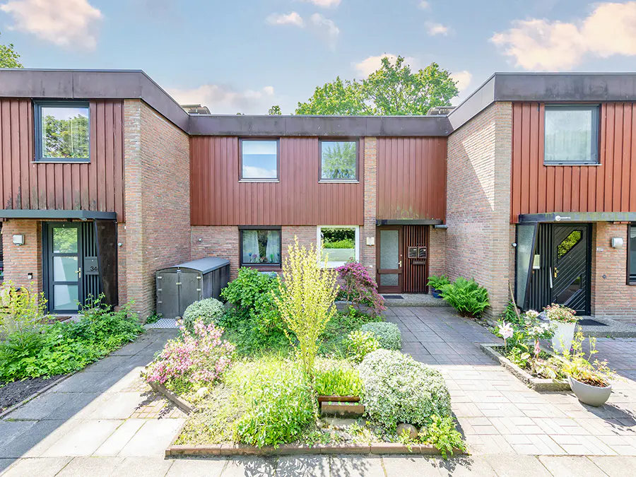 Exterior view of brick townhouses with brown siding, a paved walkway, and a garden with flowers and greenery.
