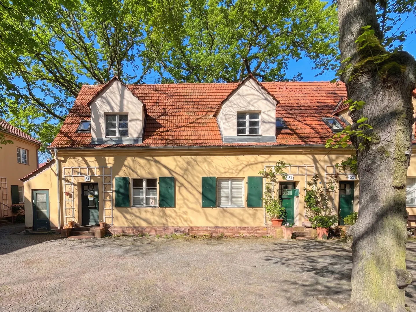 Charming yellow townhouses with red tile roofs and green shutters, framed by lush trees and a blue sky.