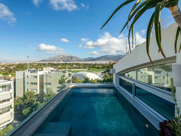 Rooftop pool with glass railing overlooks city buildings and distant mountains under a blue sky with scattered clouds.