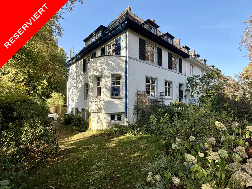 Exterior of a white, multi-story house with black shutters and a red "Reserviert" banner. Lush green lawn and trees surround the property.