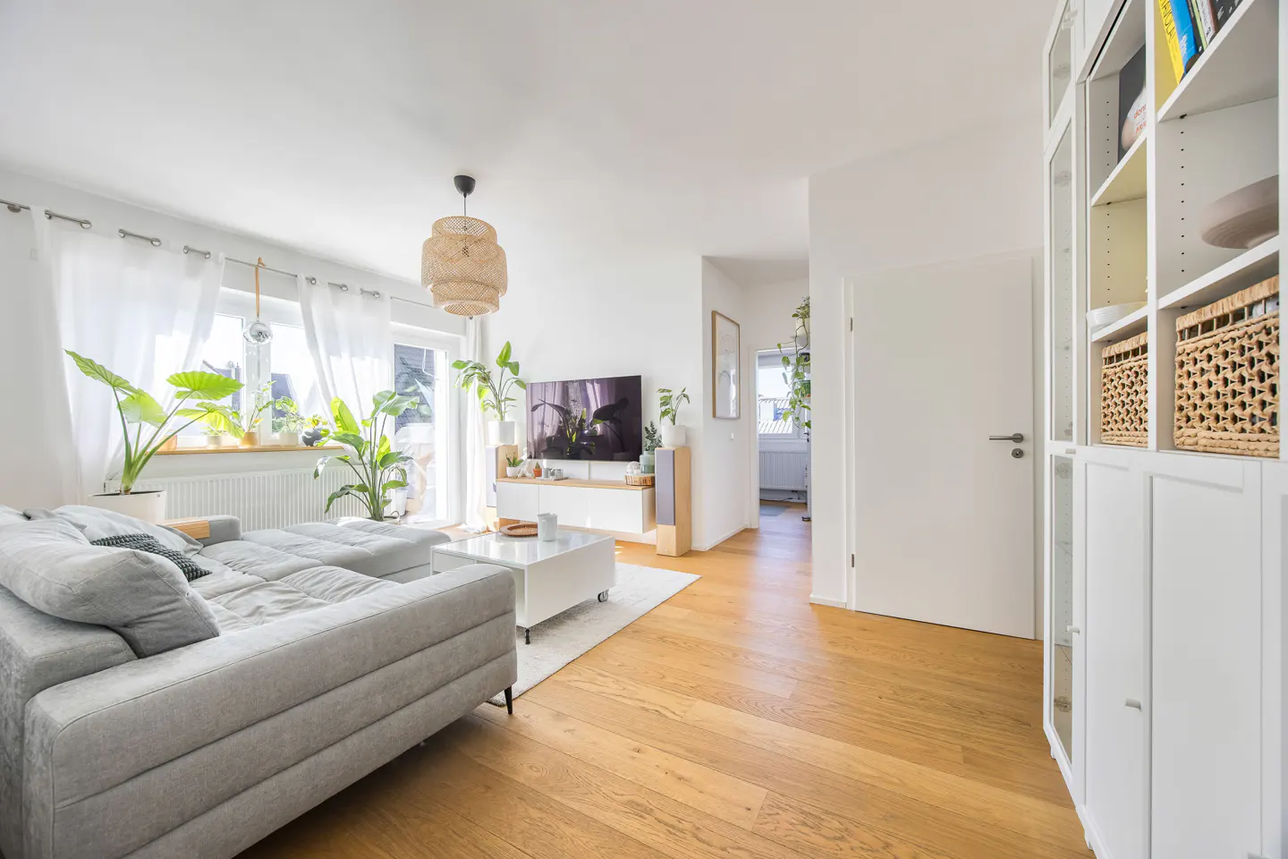 Bright living room with a gray sectional sofa, white walls, wood floors, and large windows with sheer curtains. Plants add a touch of nature.