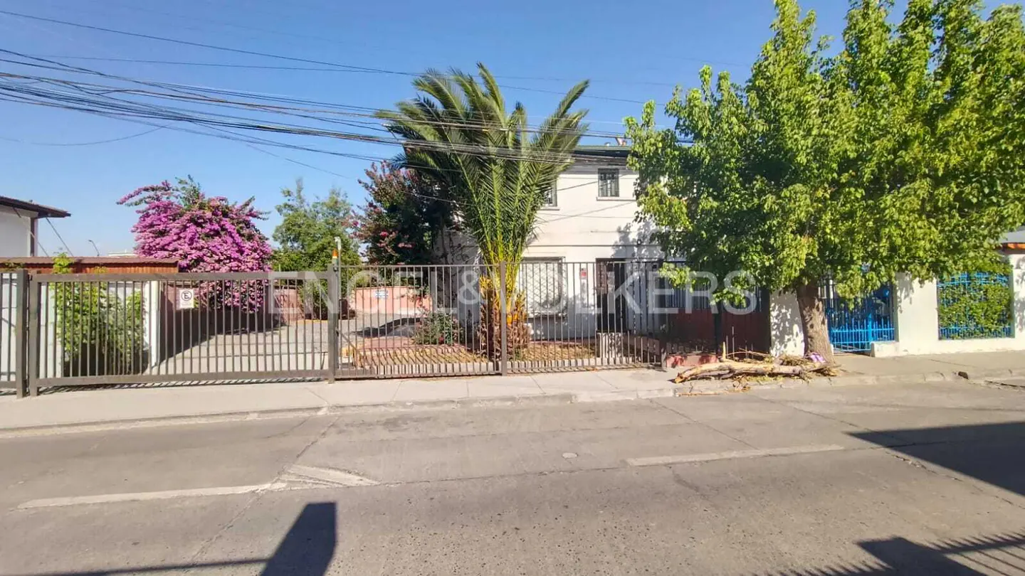 Two-story white house with a metal gate, palm tree, and green trees in front. The Engel & Volkers logo is superimposed on the image.