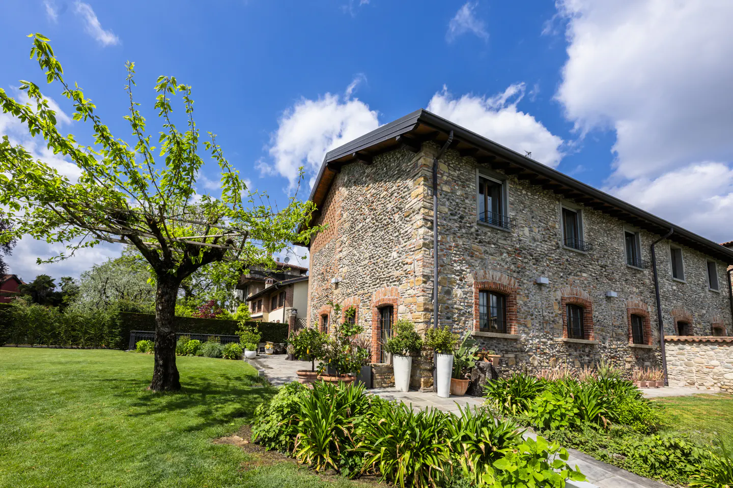 Stone house with green lawn and blue sky. The house has dark framed windows and a dark roof. A tree is on the left.
