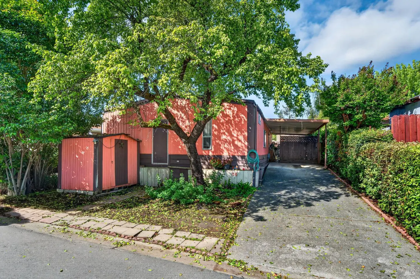 A salmon-colored single-story home with a carport and a shed, shaded by a large tree.