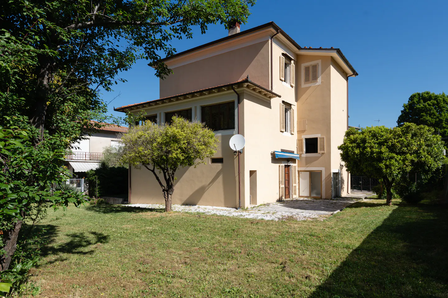 Two-story beige house with a green lawn, trees, and a blue sky in the background. A satellite dish is mounted on the side of the house.