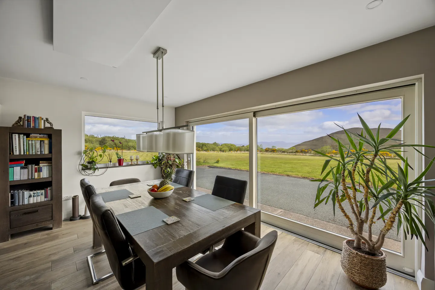 Dining room with wooden table, dark chairs, bookshelf, and large windows overlooking a scenic rural landscape.