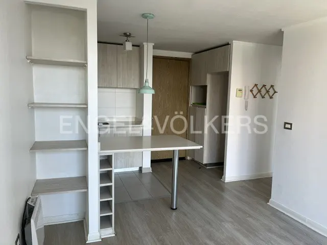 A bright apartment interior with light wood floors, white walls, and a small kitchen area with a breakfast bar.