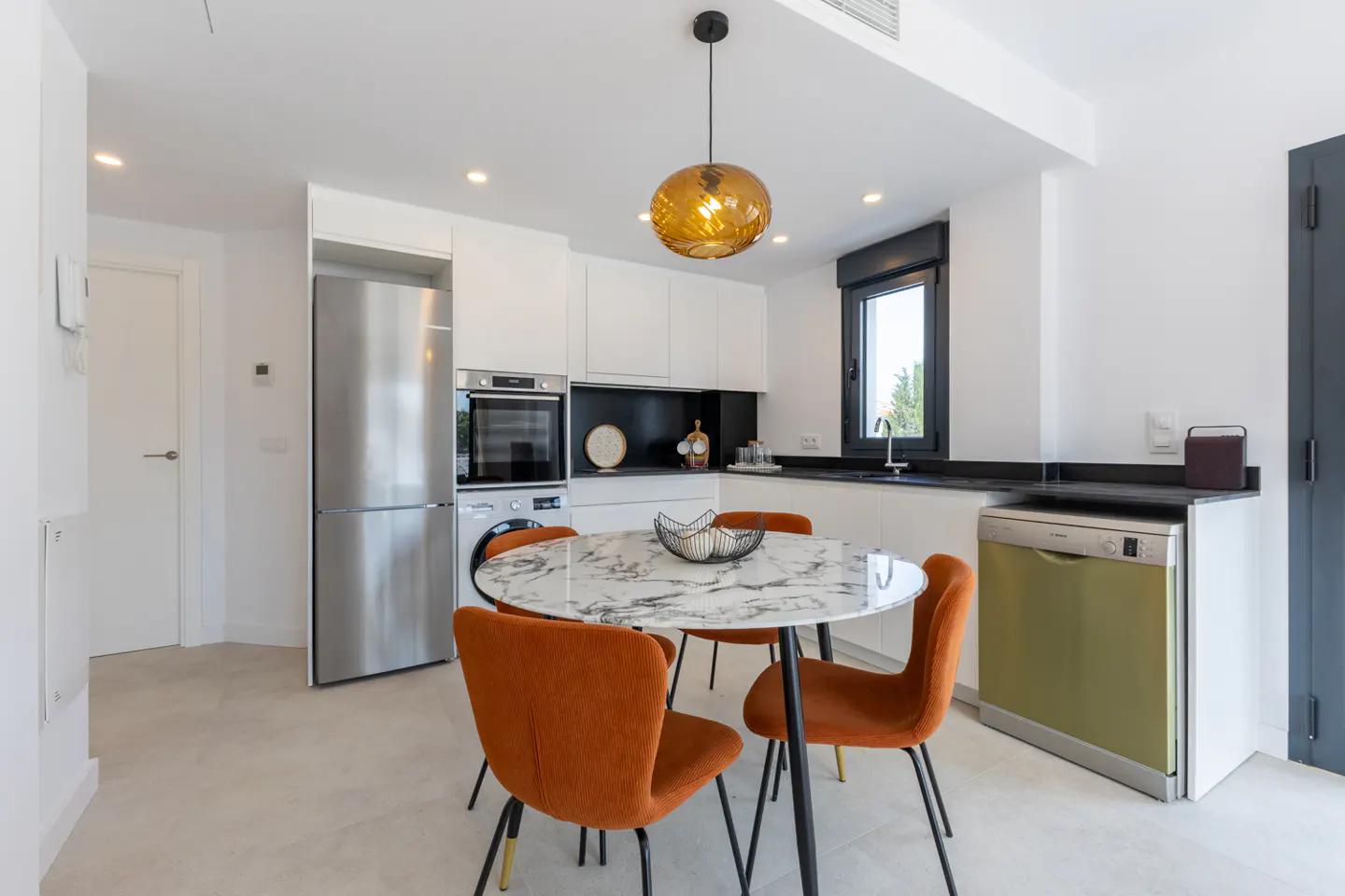 A modern kitchen with white cabinets, stainless steel appliances, and a round marble table with orange chairs. A gold pendant light hangs above the table.