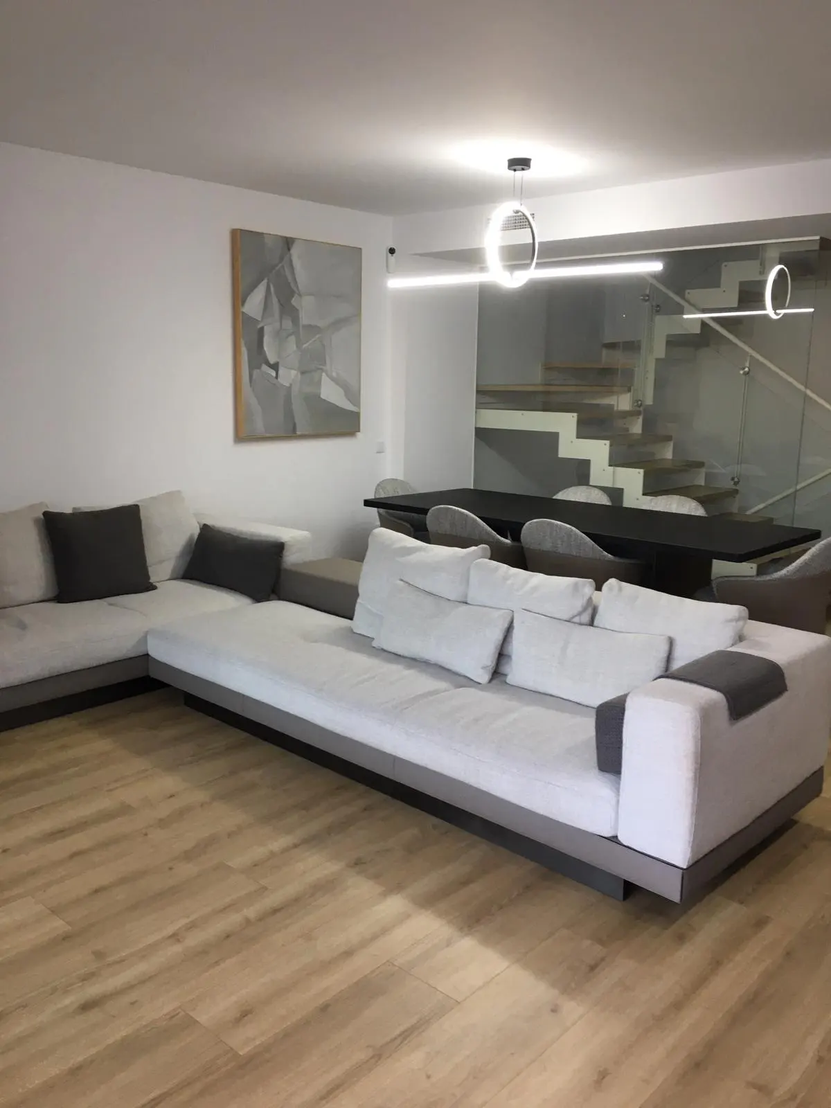 A modern living room with a large sectional sofa, wood floors, and a dining table. A painting hangs on the wall, and stairs are visible in the background.