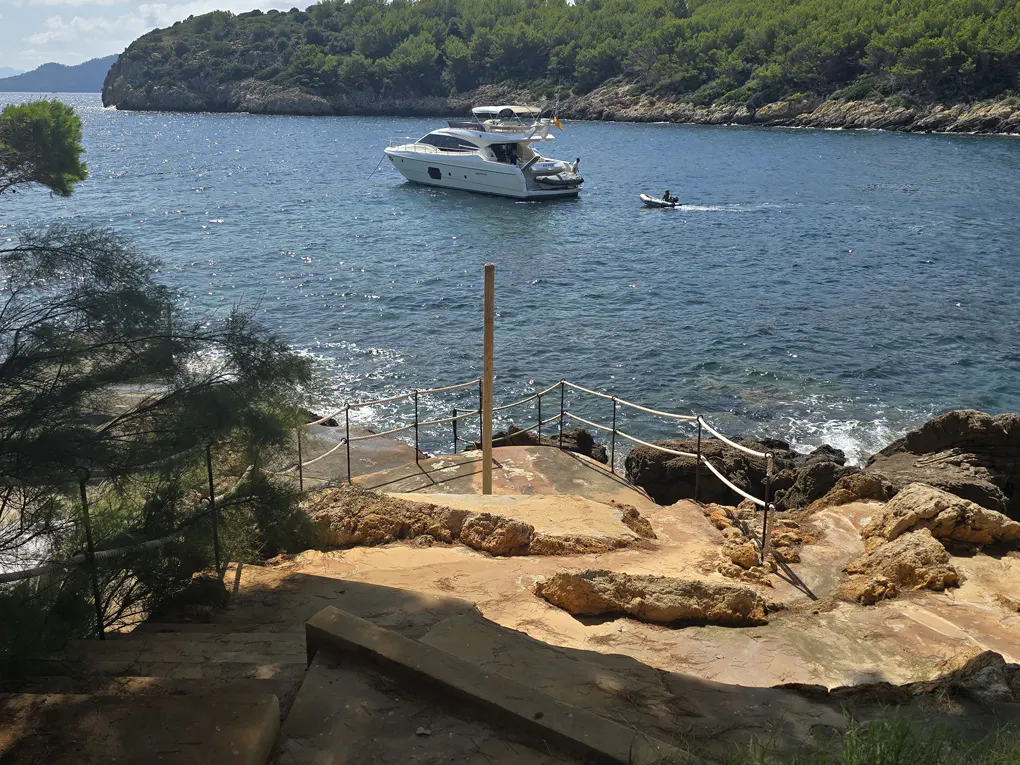 A white yacht anchored in a blue bay, viewed from a rocky, sunlit coastal path with a rope railing and pine trees.
