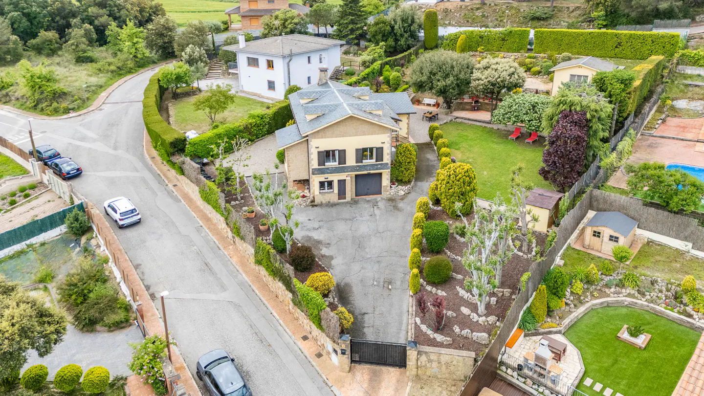 Aerial view of a tan two-story house with a gray roof, surrounded by lush green landscaping and other houses.