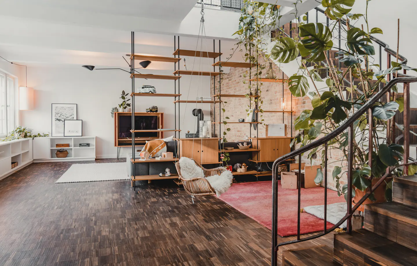 A bright living room with a wooden floor, shelves, a swing chair, and a staircase with a large green plant.