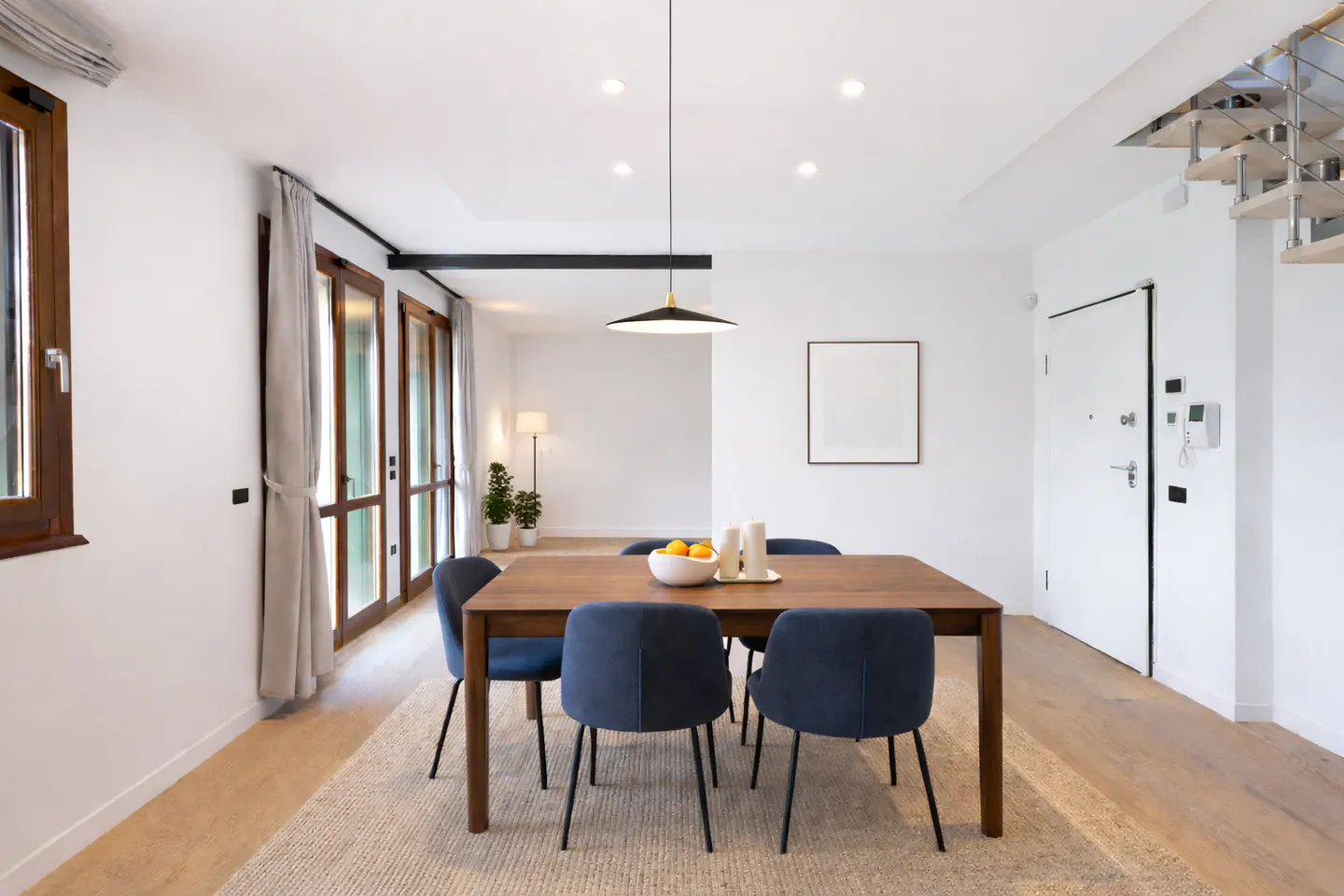 Bright dining room with a wood table, blue chairs, and a white bowl of oranges on a beige rug. White walls, wood floors, and a modern pendant light.