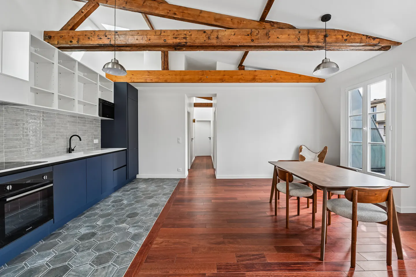 Modern kitchen with blue cabinets, grey hexagonal tile floor, wooden dining table, and exposed ceiling beams.