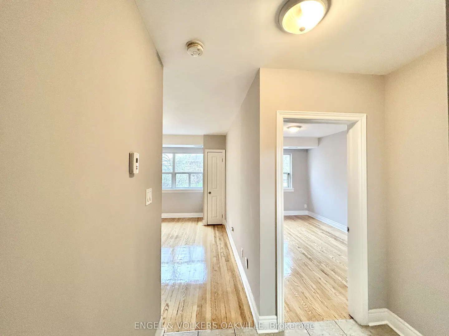 Hallway view of a home with light beige walls, hardwood floors, and white trim. Two rooms are visible through doorways.