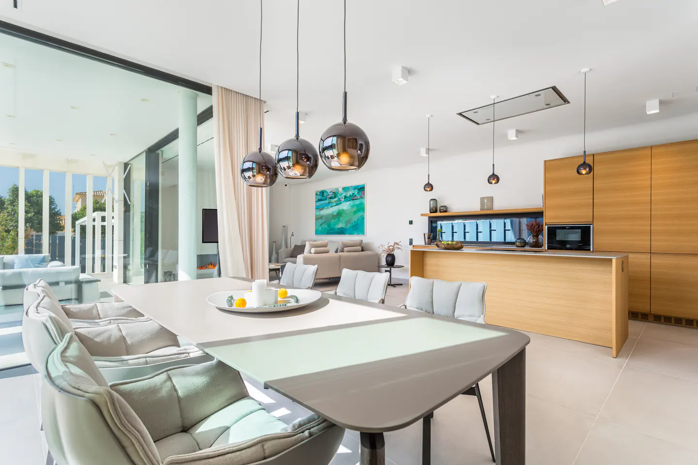 Bright, modern kitchen with a long table, upholstered chairs, and pendant lights. Large sliding glass doors open to an outdoor living space.