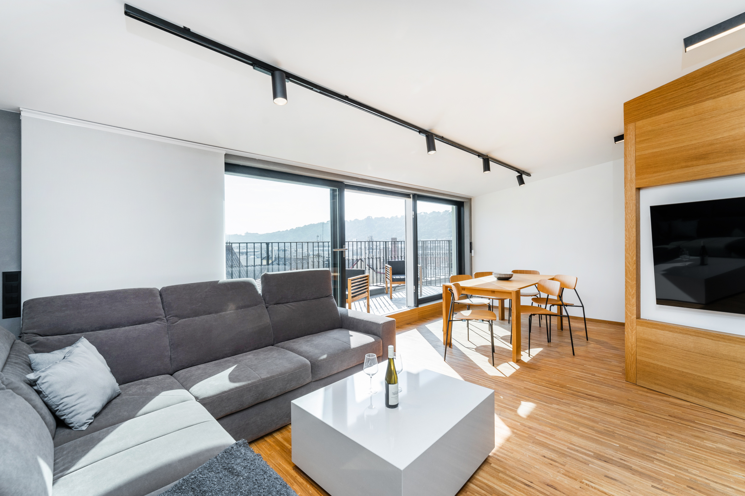 Modern living room with a grey sectional sofa, white coffee table, wooden dining set, and balcony access with city views.