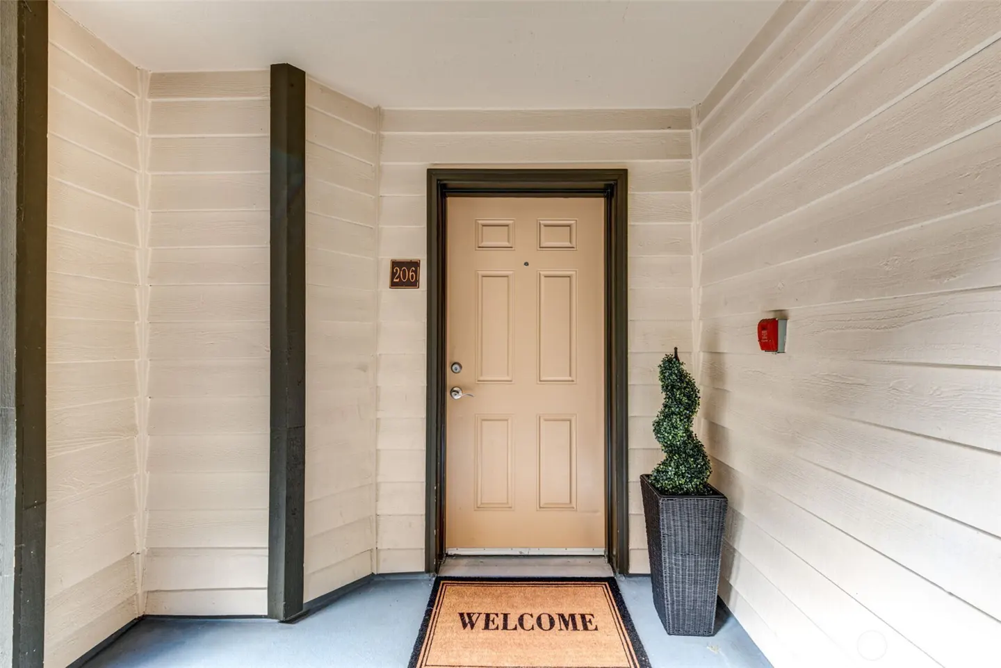 Apartment 206 entrance with a tan door, welcome mat, and potted spiral topiary. The walls are light beige with dark trim.