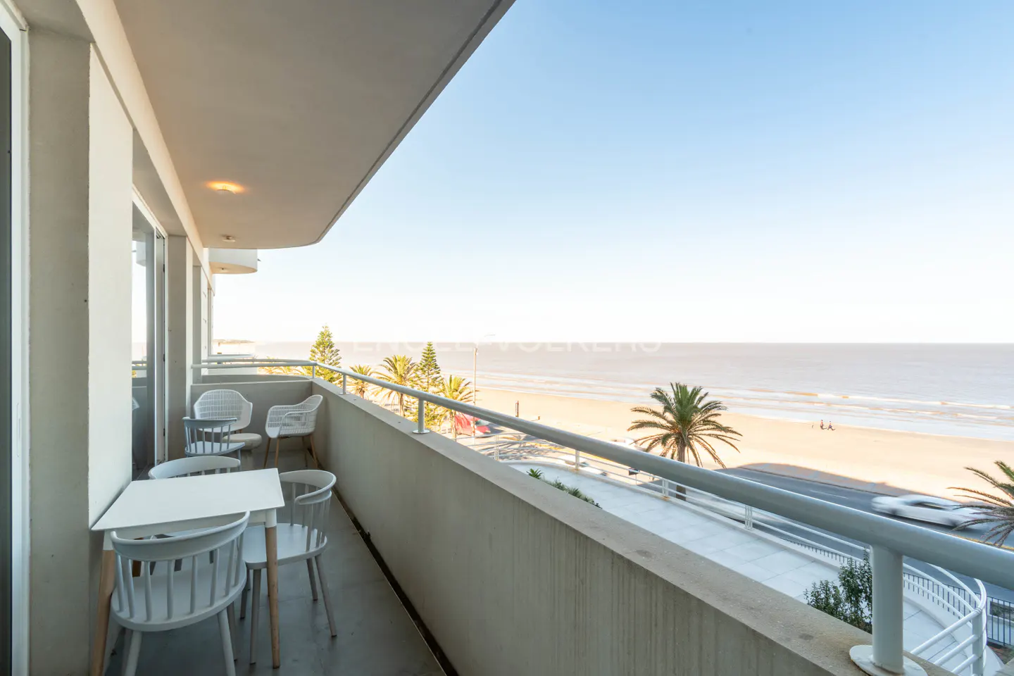 Balcony view of a beach with white table and chairs. Palm trees and ocean are visible.
