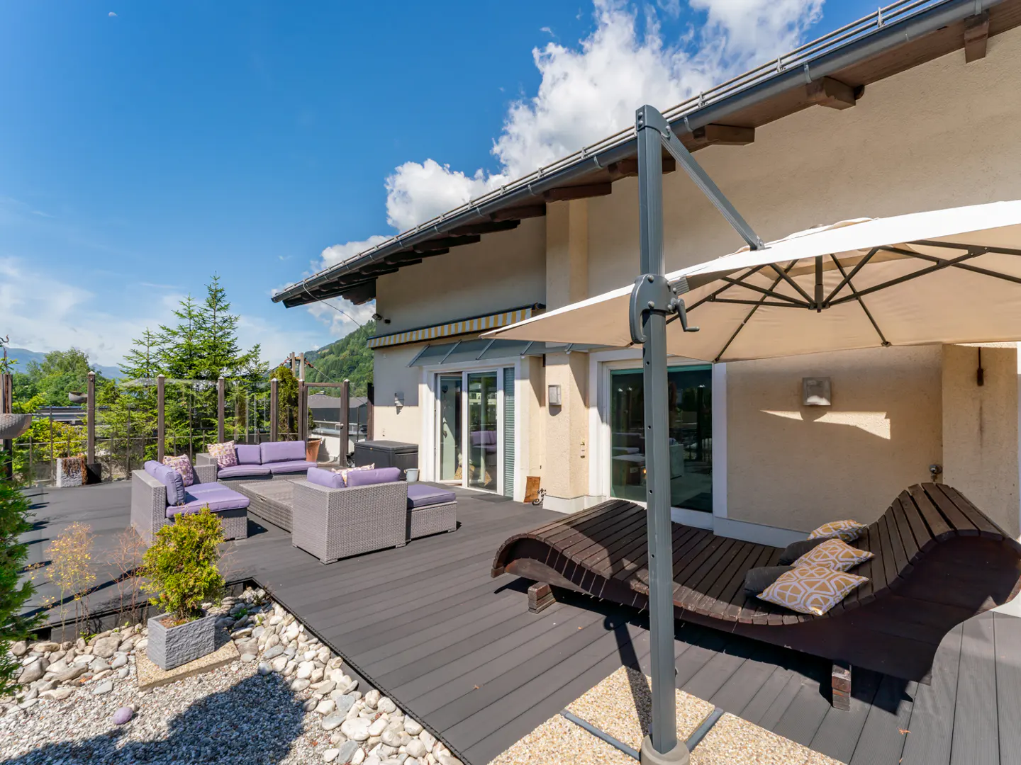 Outdoor patio with gray wicker furniture, purple cushions, and a wooden chaise lounge under a beige umbrella.