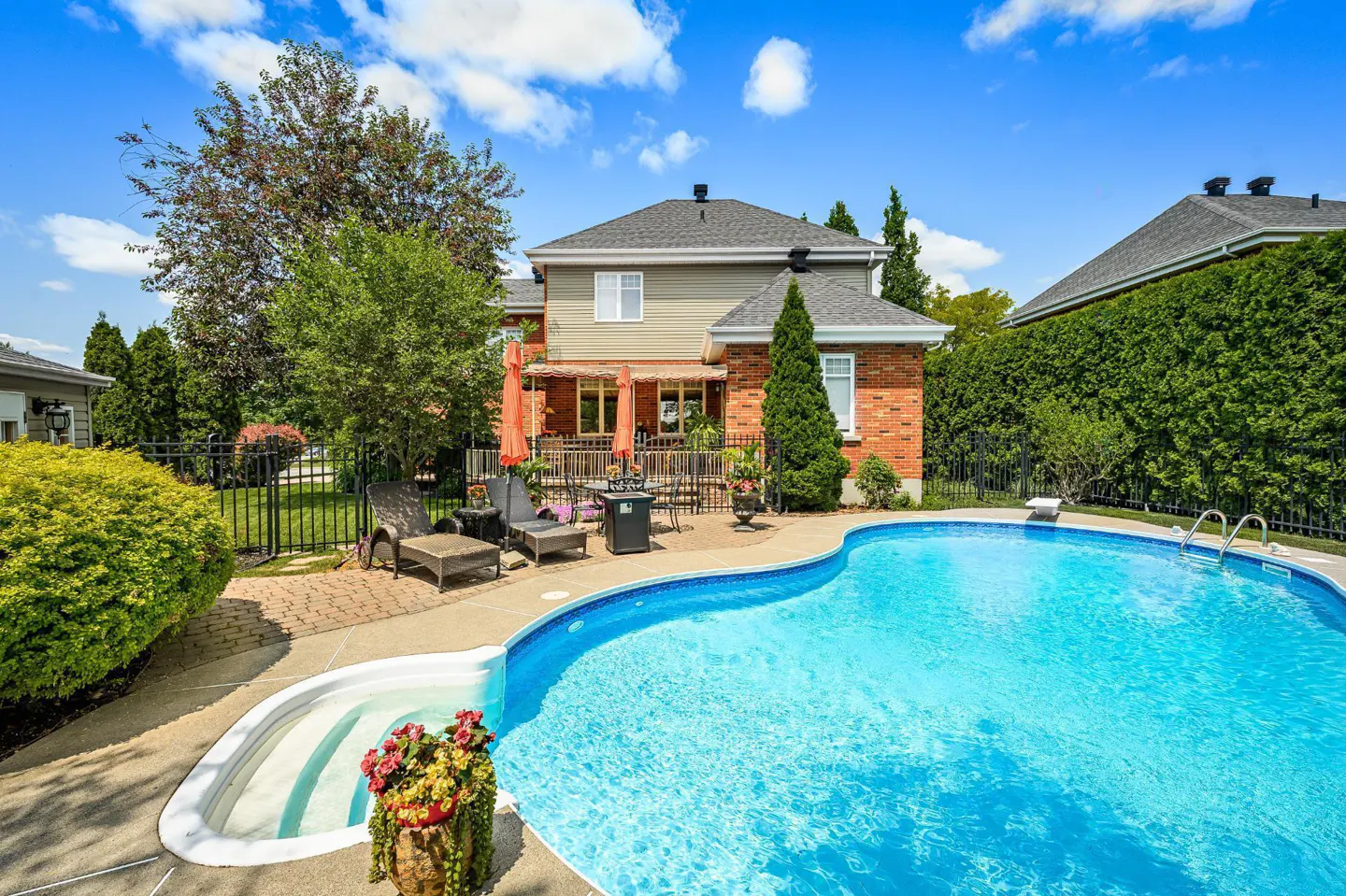 Backyard view of a two-story house with a blue in-ground pool, patio furniture, and green landscaping under a sunny sky.