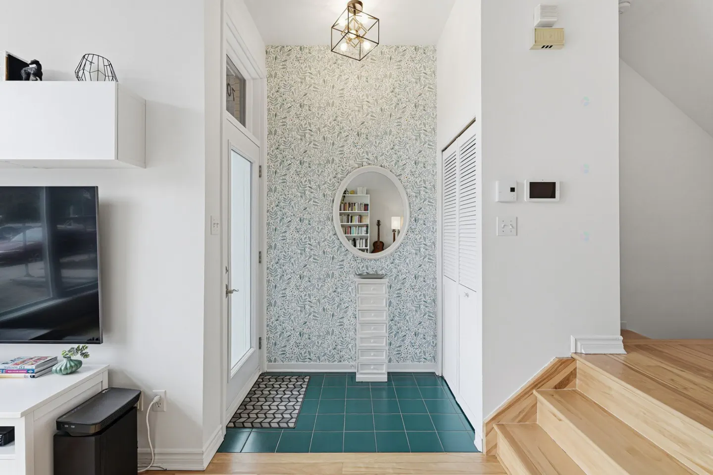 Entryway with white door, teal tile, and leaf-patterned wallpaper. An oval mirror reflects a bookshelf and guitar. A modern light fixture hangs above.