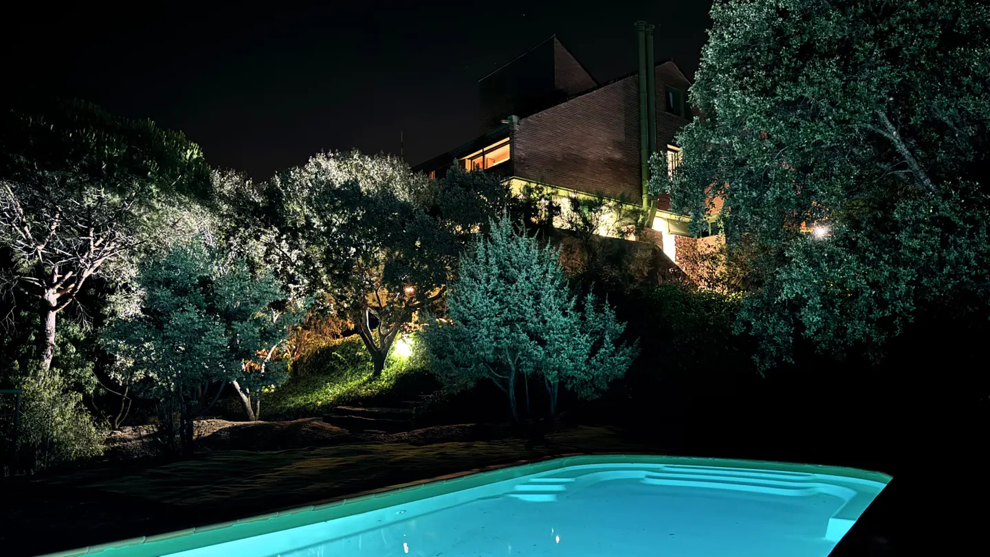 Night view of a lit pool in front of a brick house, surrounded by trees and greenery.