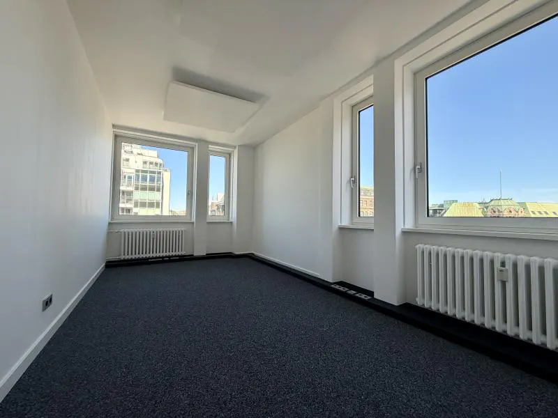 Bright, empty office space with white walls, blue carpet, and large windows showing city views. Radiators are under the windows.