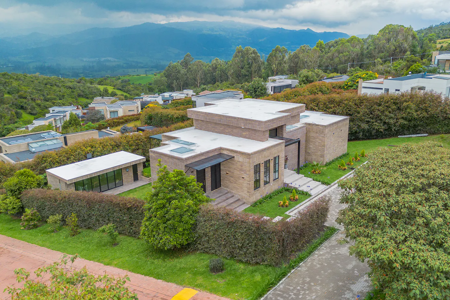 Aerial view of a modern, tan brick house with a flat white roof, green lawn, and mountain backdrop.