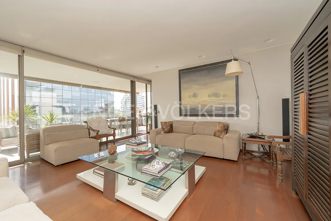 Living room with beige sofas, glass coffee table, and wood floors. Balcony with city views visible through sliding glass doors.