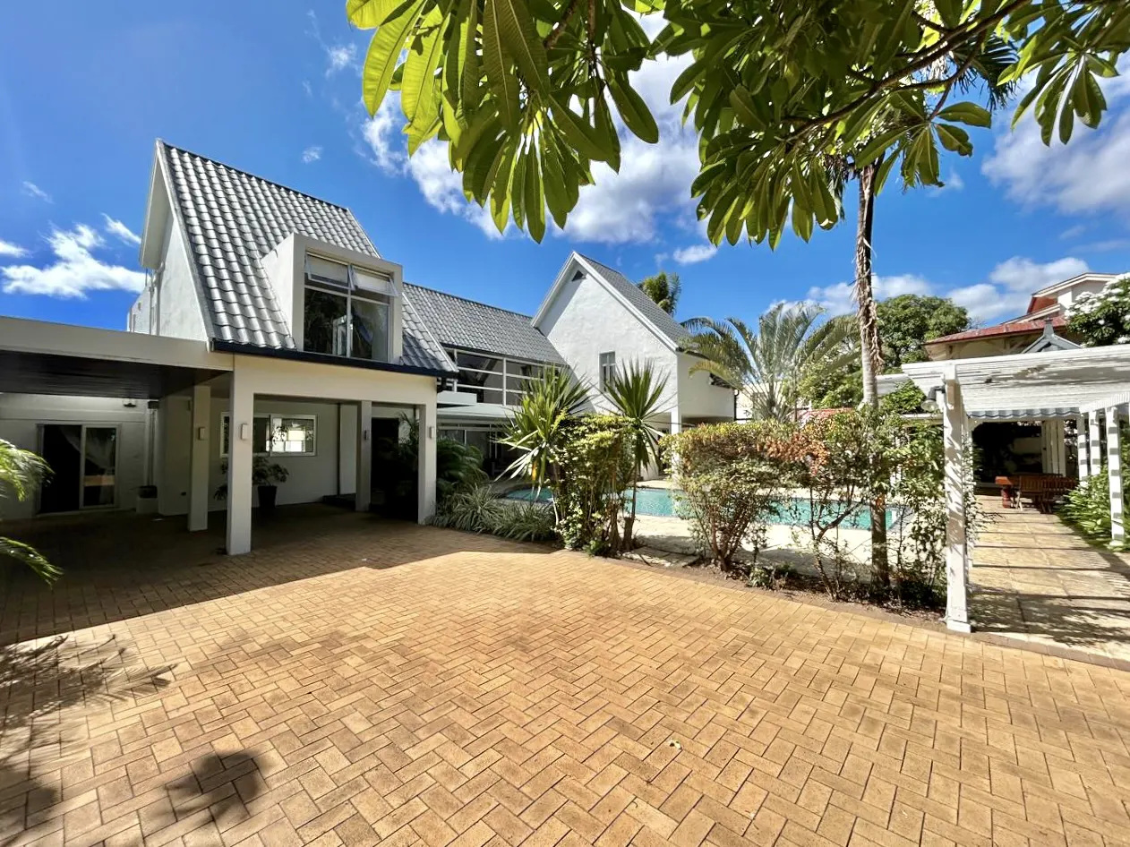 Exterior view of a modern white house with a gray roof, brick driveway, pool, and lush tropical landscaping.