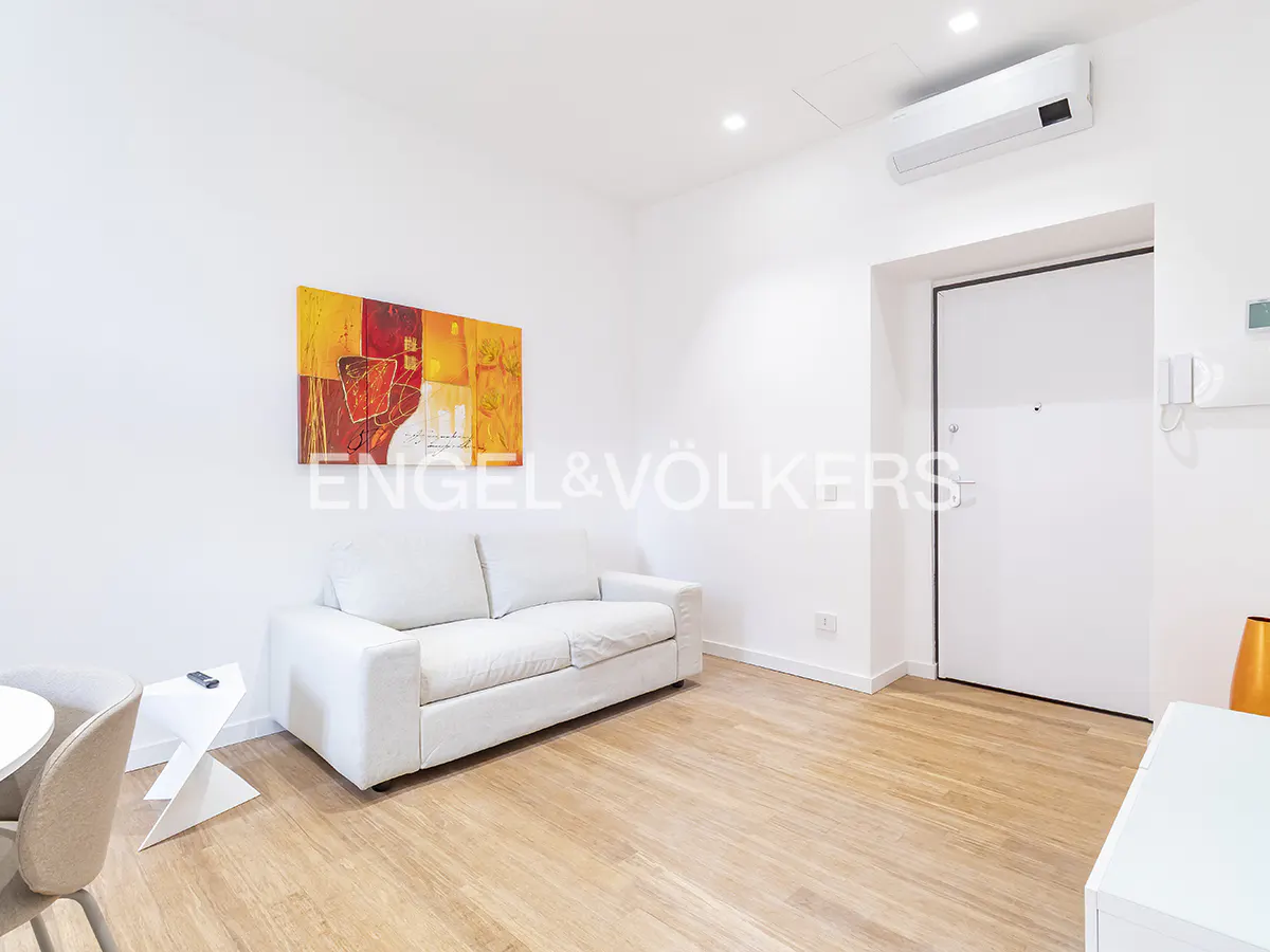 Bright living room with white walls, wood floors, and a white sofa under an abstract painting. Engel & Völkers logo visible.