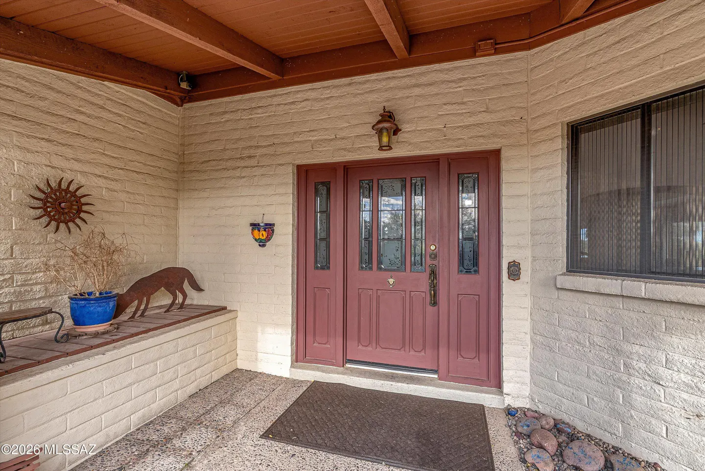 Front entrance of a home with a red door, brick walls, and a brown ceiling. A metal sun and coyote statue decorate the porch.