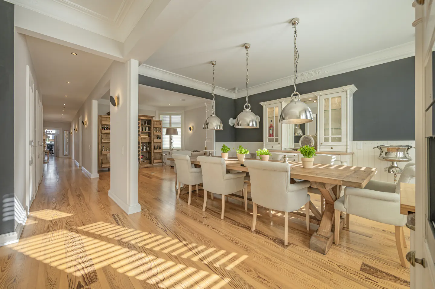 Bright dining room with a long wooden table, beige chairs, and silver pendant lights. A hallway with wood floors is visible to the left.