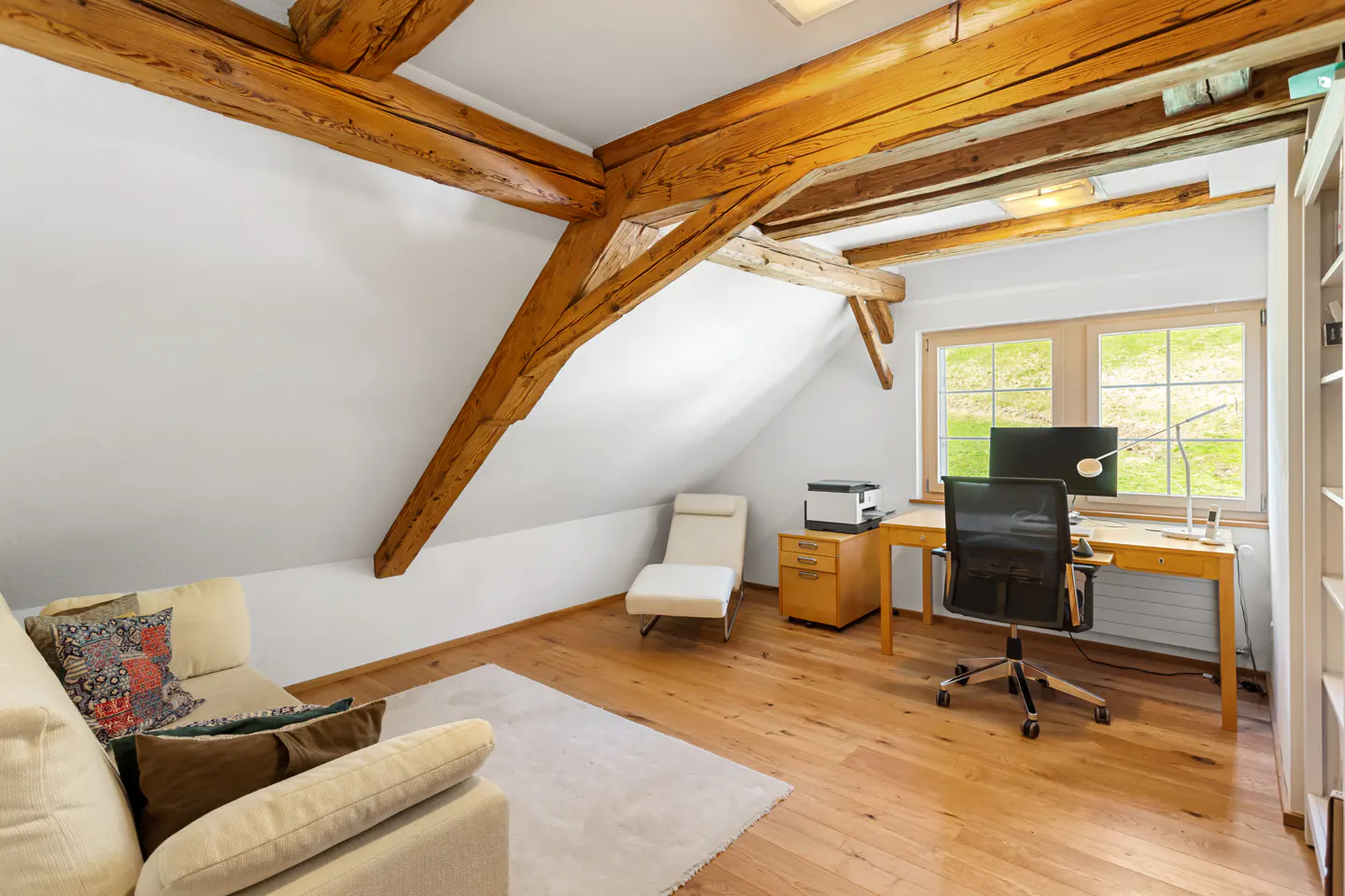 Attic room with exposed wooden beams, white walls, and hardwood floors. A desk, chair, and sofa create a cozy workspace.