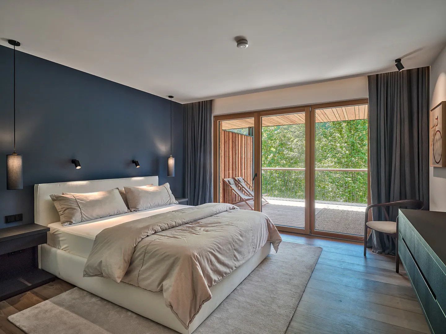 Modern bedroom with a navy accent wall, white bed, and large glass doors opening to a balcony with forest views.