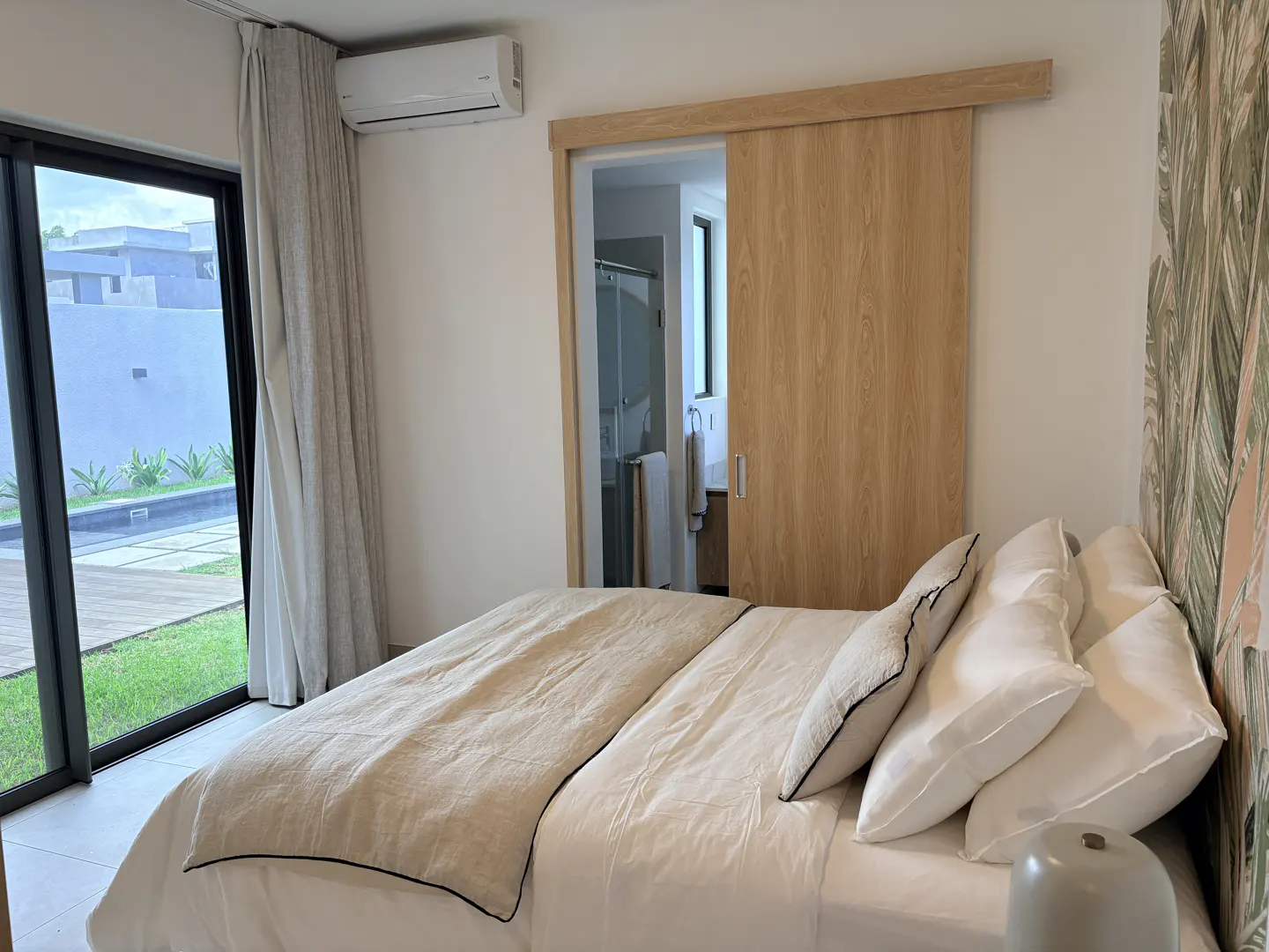A bright bedroom with a large bed, sliding wood door to a bathroom, and a glass door to a patio and lawn.