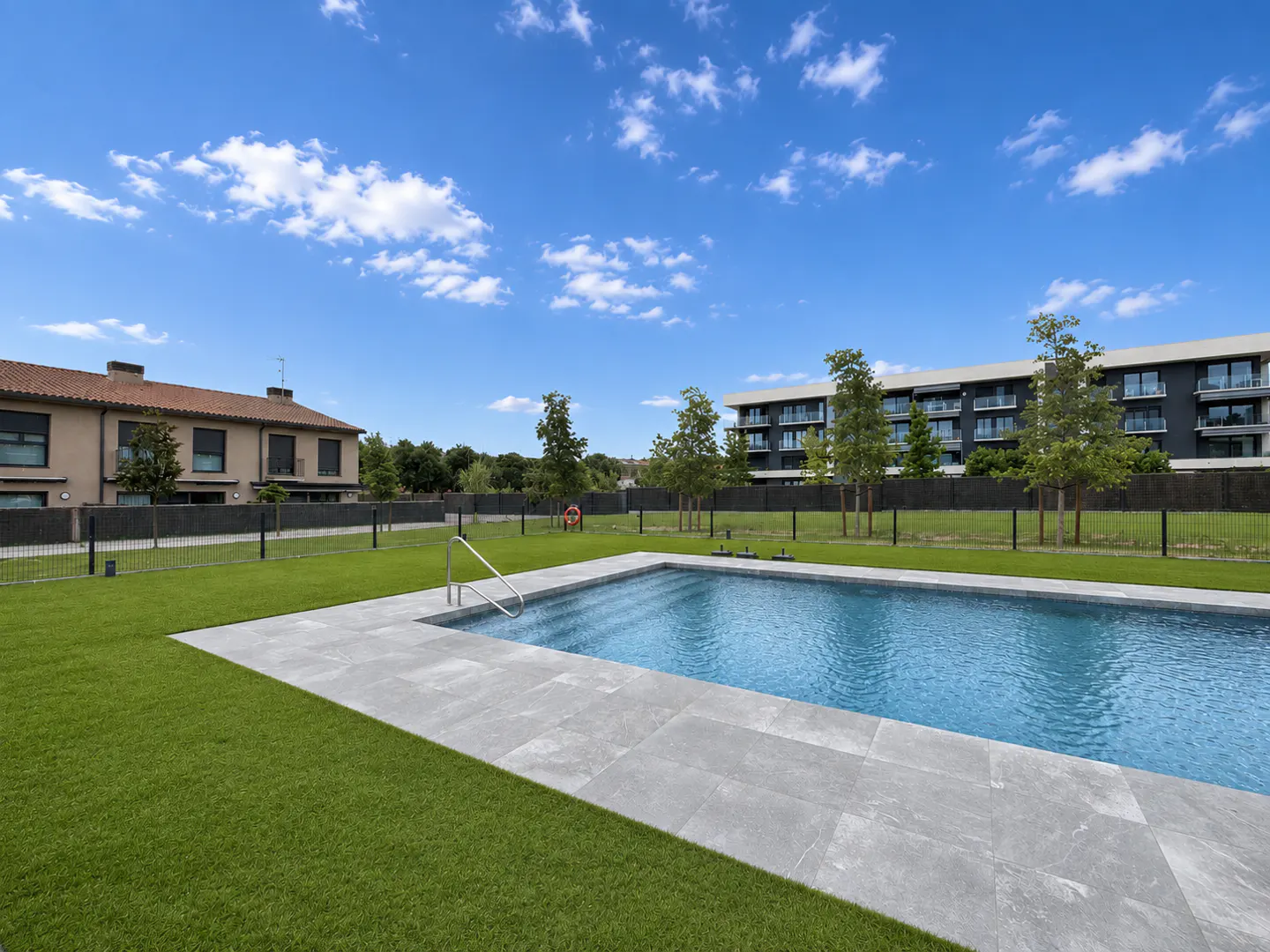A rectangular swimming pool with a grey stone deck set in a green lawn, with modern apartment buildings in the background.