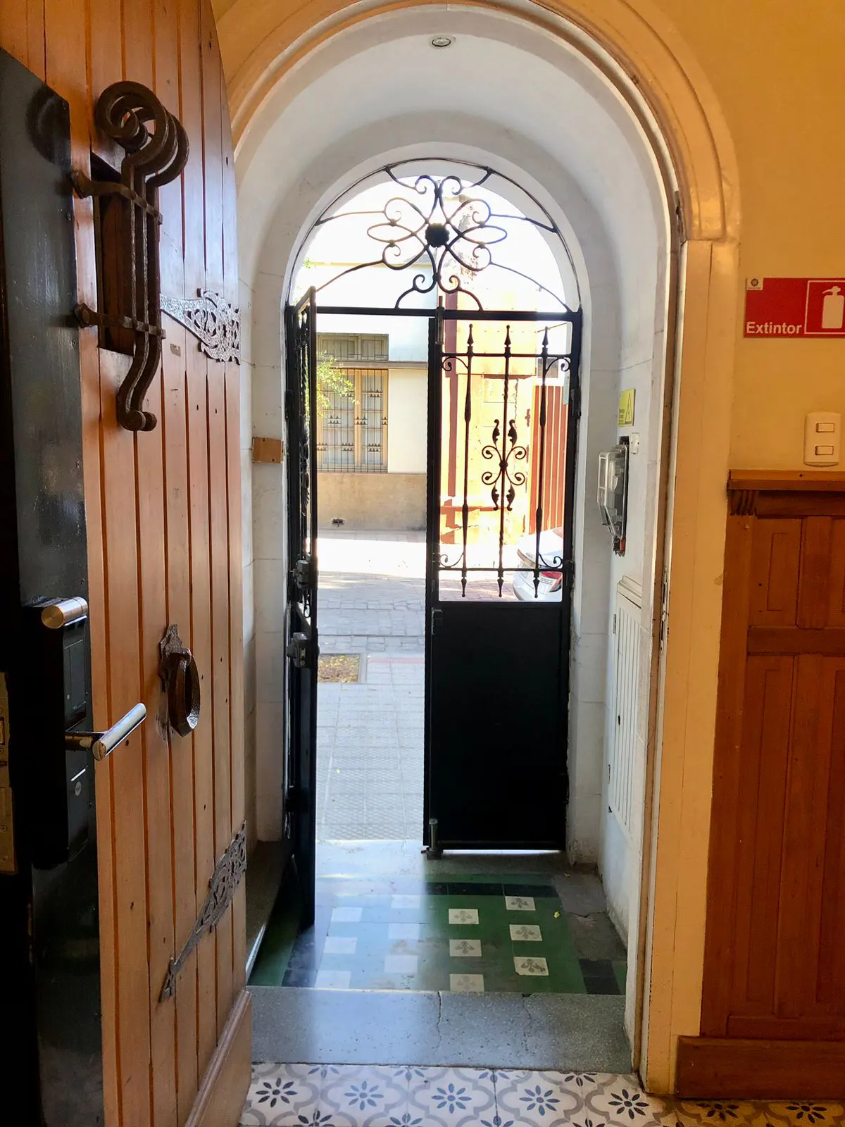 Open wooden door with ornate iron hardware, leading to an arched entryway with a black iron gate and view to the outside.