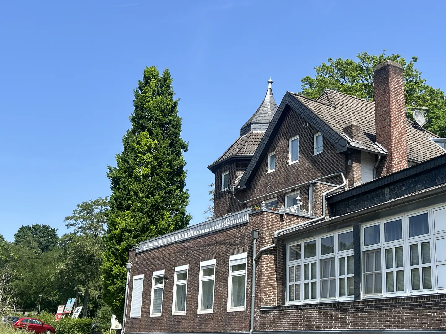 Exterior view of a brown brick building with white windows under a clear blue sky. A tall green tree stands beside the building.