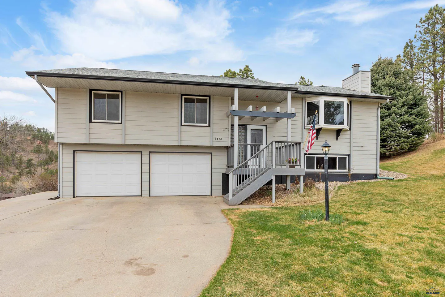Two-story house with gray siding, white garage doors, and a gray staircase leading to the front door. An American flag hangs near the entrance.