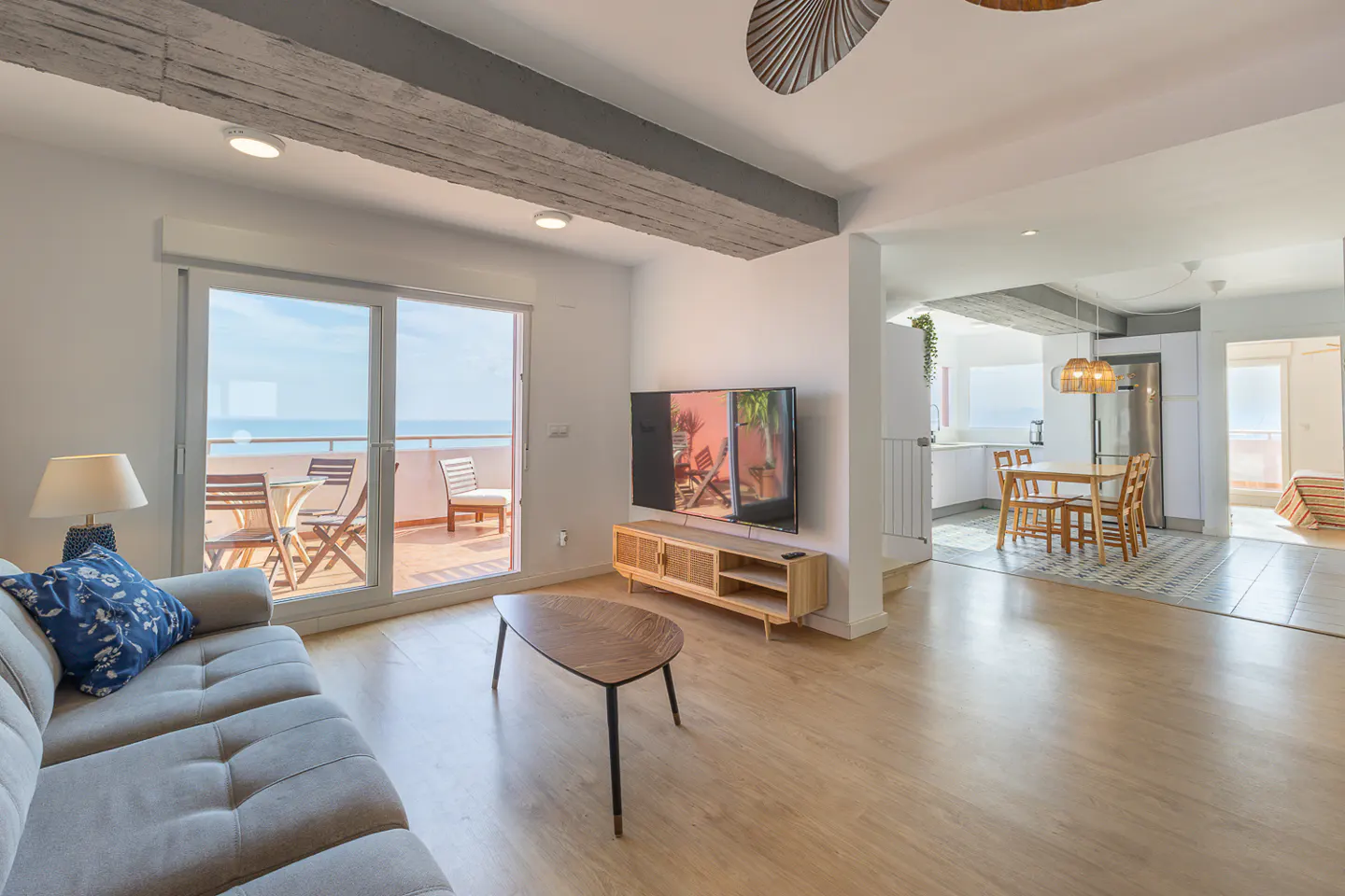 Bright, open-concept living space with a gray sofa, wood floors, and a balcony overlooking the ocean. A kitchen and dining area are visible in the background.