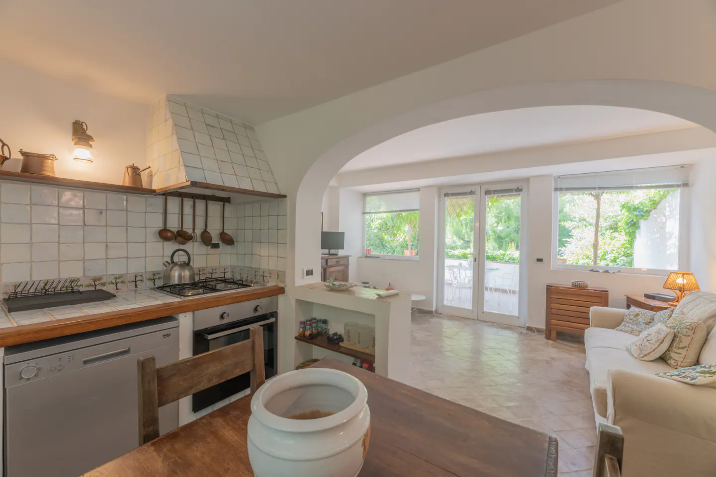 Open-concept kitchen and living room with white walls, tile floors, and wood accents. A white sofa faces windows with a garden view.
